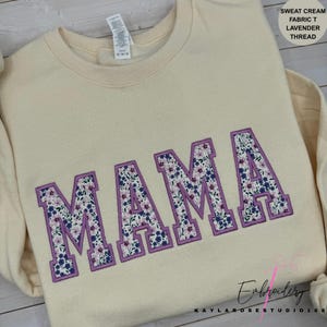 May include: Cream-colored sweatshirt with the embroidered word "MAMA" in large, floral-filled letters outlined in lavender. The floral pattern includes blue and pink flowers. The text "SWEAT CREAM FABRIC T LAVENDER THREAD" is in the upper right corner.