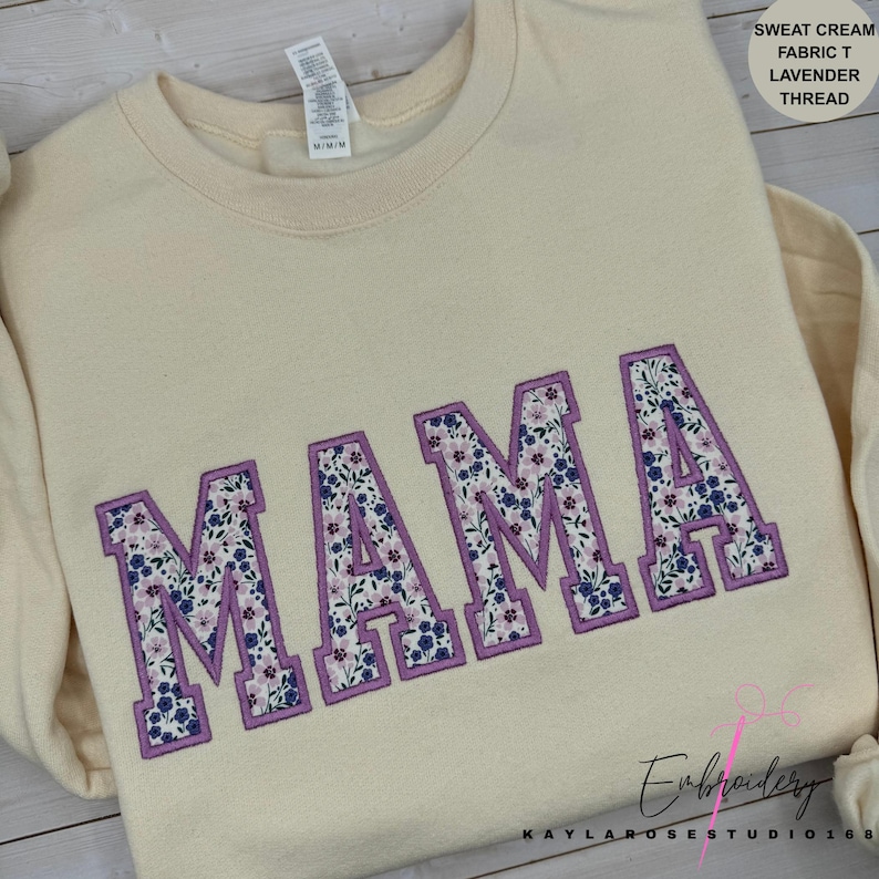May include: Cream-colored sweatshirt with the embroidered word "MAMA" in large, floral-filled letters outlined in lavender. The floral pattern includes blue and pink flowers. The text "SWEAT CREAM FABRIC T LAVENDER THREAD" is in the upper right corner.