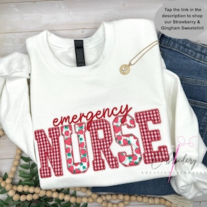 Op de afbeelding: Een wit sweatshirt met lange mouwen met de rode tekst emergency en het woord NURSE in een rood-wit gingham patroon gevuld met aardbei-illustraties. Een goudkleurige hangerketting rust op de stof.