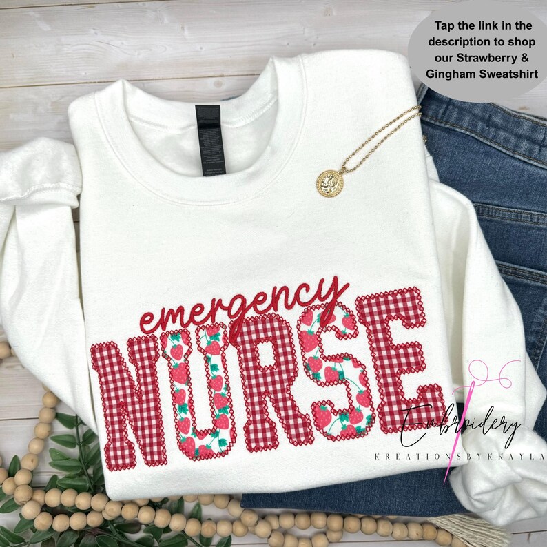 Op de afbeelding: Een wit sweatshirt met lange mouwen met de rode tekst emergency en het woord NURSE in een rood-wit gingham patroon gevuld met aardbei-illustraties. Een goudkleurige hangerketting rust op de stof.