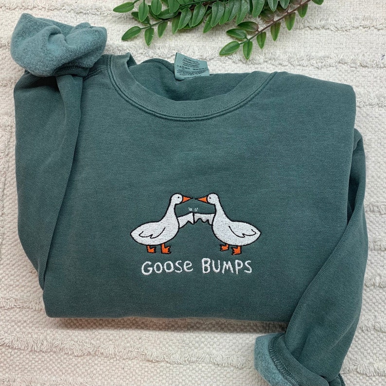 K&ouml;nnte beinhalten: Ein dunkelgr&uuml;ner Pullover mit einem wei&szlig;en gestickten Design aus zwei sich gegen&uuml;berstehenden G&auml;nsen. Die G&auml;nse halten ein St&uuml;ck Papier mit dem Text "Goose Bumps" fest.
