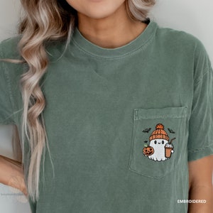 May include: Olive green t-shirt with an embroidered pocket design. The design features a cartoon ghost wearing an orange beanie, holding a pumpkin and a coffee drink. The word "EMBROIDERED" is printed on the bottom right.