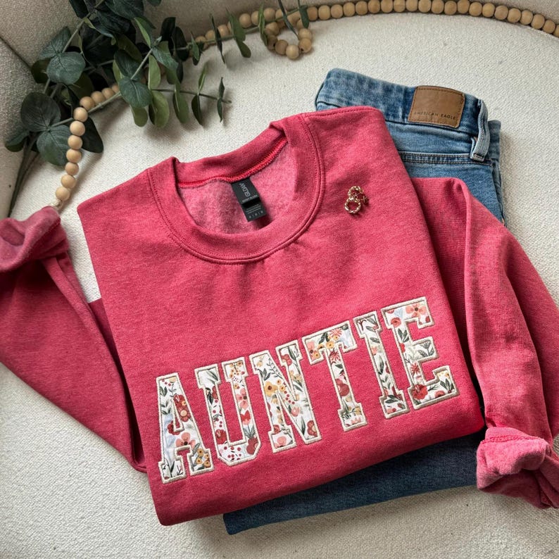 May include: A red sweatshirt with the word "AUNTIE" in floral letters. A small gold brooch is pinned near the collar. Blue jeans are folded beneath the sweatshirt. The sweatshirt has a crew neck and long sleeves.