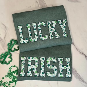 May include: Two green t-shirts with the words "LUCKY" and "IRISH" in white letters. The letters are filled with a pattern of green shamrocks.