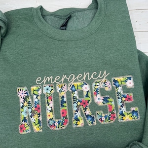 Op de afbeelding: Een gem&ecirc;leerd bosgroen sweatshirt met ronde hals voorzien van het woord "emergency" in een lichtbeige schrijfletter boven het woord "NURSE" in grote blokletters gevuld met een kleurrijk bloemenpatroon op een donkerblauwe achtergrond.