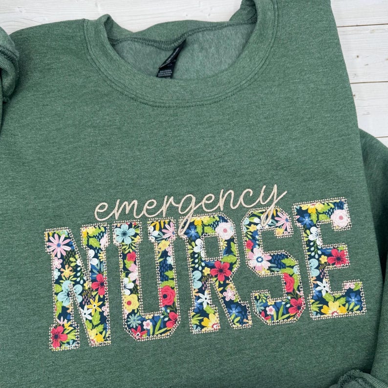 Op de afbeelding: Een gem&ecirc;leerd bosgroen sweatshirt met ronde hals voorzien van het woord "emergency" in een lichtbeige schrijfletter boven het woord "NURSE" in grote blokletters gevuld met een kleurrijk bloemenpatroon op een donkerblauwe achtergrond.