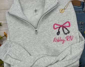 Può includere: Una felpa grigio chiaro con mezza zip, caratterizzata da uno stetoscopio rosa ricamato a forma di fiocco e dalla scritta Ashley RN in corsivo rosa. Una collana con ciondolo circolare dorato poggia sul tessuto vicino al colletto.