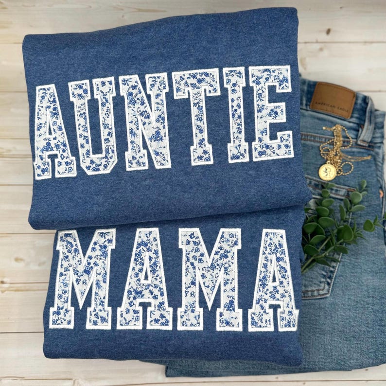 May include: Two blue sweatshirts with white floral lettering that reads "AUNTIE" and "MAMA". The sweatshirts are folded and placed next to a pair of blue jeans with a gold necklace and a sprig of greenery.