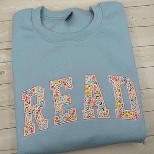 May include: A pale blue sweatshirt featuring the embroidered word "READ". The letters are filled with a floral design in pink, yellow, and blue, and are outlined in pink.
