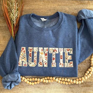 May include: Auntie sweatshirt in a denim blue color. The word "AUNTIE" is displayed across the front in a floral design with red, orange, and yellow flowers. The sweatshirt has a crew neck and long sleeves.