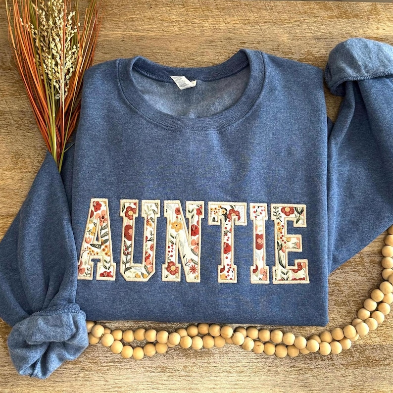 May include: Auntie sweatshirt in a denim blue color. The word "AUNTIE" is displayed across the front in a floral design with red, orange, and yellow flowers. The sweatshirt has a crew neck and long sleeves.