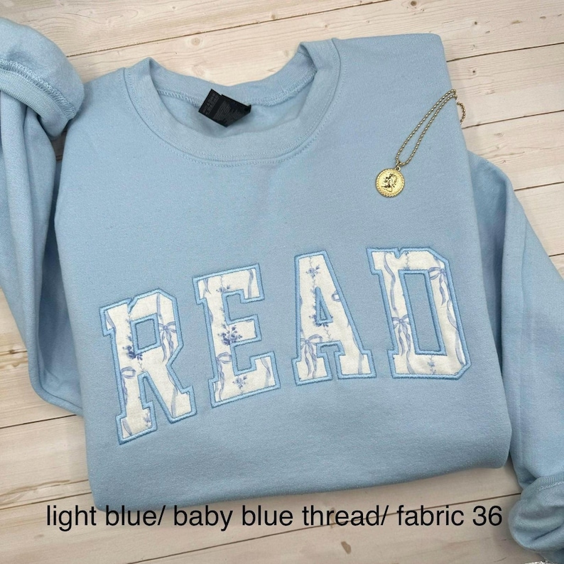 May include: A pale blue sweatshirt featuring the word "READ" embroidered in white with a floral design. A gold-coloured necklace with a pendant is draped over the sweatshirt. The text "light blue/ baby blue thread/ fabric 36" is at the bottom.