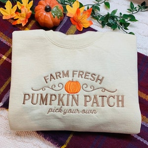 May include: A light beige sweatshirt with the text "Farm Fresh Pumpkin Patch pick your own" embroidered in brown. A small orange pumpkin is embroidered in the centre of the text.