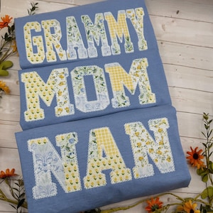 May include: Three light blue t-shirts with floral patchwork letters spelling "GRAMMY", "MOM", and "NAN". The letters are in yellow, white, and blue floral patterns, with a white lace trim. The shirts are displayed on a wooden surface.
