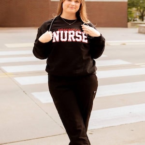 Op de afbeelding: Een zwart sweatshirt met ronde hals en de tekst "emergency NURSE" in witte en rode letters op de borst, gecombineerd met een zwarte werkbroek en witte sportschoenen. Een stethoscoop hangt om de nek.