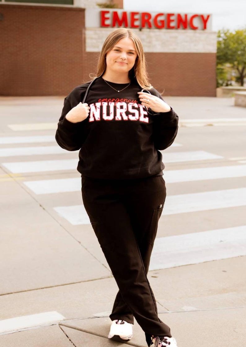 Op de afbeelding: Een zwart sweatshirt met ronde hals en de tekst "emergency NURSE" in witte en rode letters op de borst, gecombineerd met een zwarte werkbroek en witte sportschoenen. Een stethoscoop hangt om de nek.