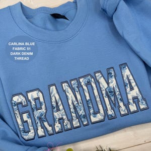 May include: A light blue sweatshirt with the word "GRANDMA" embroidered across the front in a floral pattern. The text "CARLINA BLUE FABRIC 51 DARK DENIM THREAD" is printed on the upper left side of the sweatshirt.