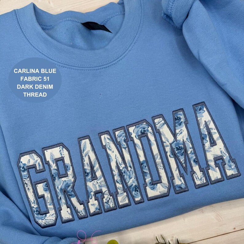May include: A light blue sweatshirt with the word "GRANDMA" embroidered across the front in a floral pattern. The text "CARLINA BLUE FABRIC 51 DARK DENIM THREAD" is printed on the upper left side of the sweatshirt.