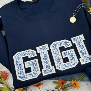 以下が含まれることがあります： ネイビーブルーのスウェットシャツ。白い縁取りと青い花柄で「GIGI」という名前が大きく書かれています。金のペンダントネックレスが見えます。スウェットシャツは折りたたまれており、隅には花があります。