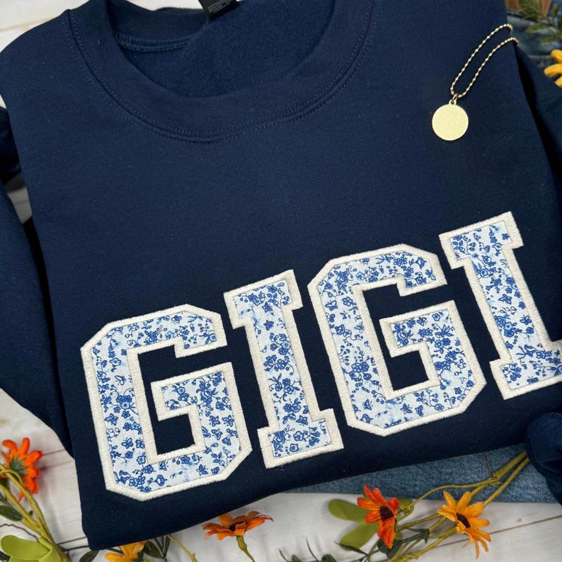 以下が含まれることがあります： ネイビーブルーのスウェットシャツ。白い縁取りと青い花柄で「GIGI」という名前が大きく書かれています。金のペンダントネックレスが見えます。スウェットシャツは折りたたまれており、隅には花があります。