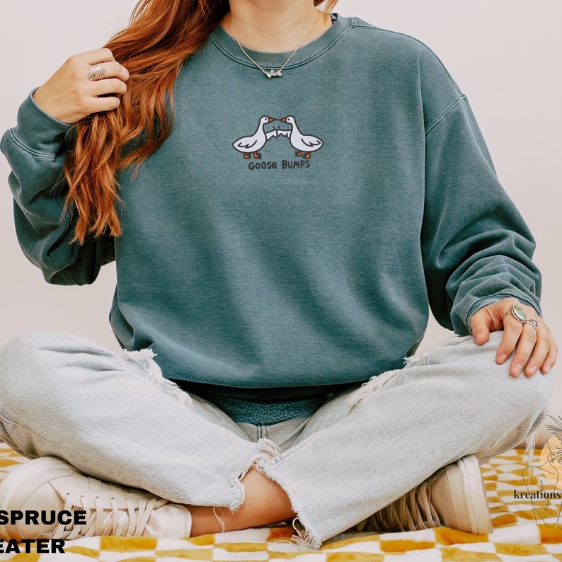 K&ouml;nnte beinhalten: Ein blaugr&uuml;ner Pullover mit einem wei&szlig;en gestickten Design aus zwei sich gegen&uuml;berstehenden G&auml;nsen mit dem Text "Goose Bumps" darunter.