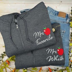May include: Two gray zip-up hoodies with white cursive text and a red apple graphic. One says "Mrs. Fucarile" and the other says "Mrs. White." Light blue denim jeans are visible in the background.