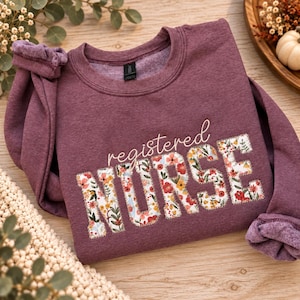 May include: A heathered maroon crewneck sweatshirt featuring the text registered NURSE in a cursive and block font. The word NURSE is filled with a colorful floral pattern of red, orange, yellow, and green flowers on a white background.