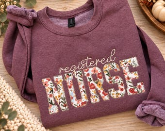 May include: A heathered maroon crewneck sweatshirt featuring the text registered NURSE in a cursive and block font. The word NURSE is filled with a colorful floral pattern of red, orange, yellow, and green flowers on a white background.