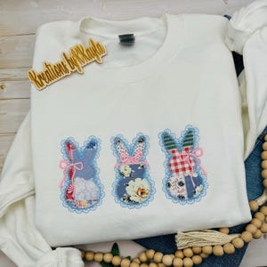 May include: White sweatshirt with three appliqued bunny designs. Each bunny is a different pattern, with blue lace trim and pink bows. The wooden tag says "Kreations by Kayla".