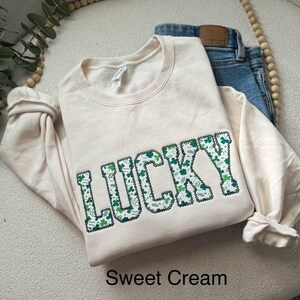 May include: A cream-colored sweatshirt with the word "LUCKY" spelled out in green shamrock-patterned letters. The text is on a cream-colored sweatshirt. The text "Sweet Cream" is written below the sweatshirt.