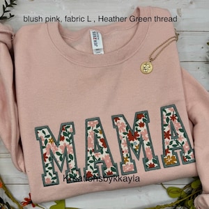 May include: Blush pink sweatshirt with the word "MAMA" embroidered in a floral pattern. The letters are outlined in green thread. A gold necklace with a pendant is draped on the sweatshirt.