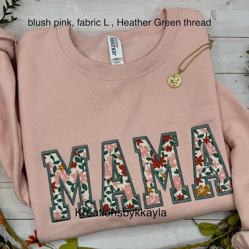 May include: Blush pink sweatshirt with the word "MAMA" embroidered in a floral pattern. The letters are outlined in green thread. A gold necklace with a pendant is draped on the sweatshirt.