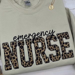 May include: A beige crew neck sweatshirt with the words "emergency NURSE" in black script and leopard print letters. A gold pendant necklace is draped over the sweatshirt.