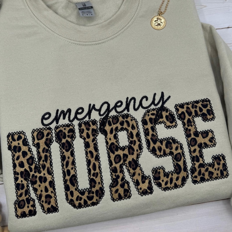 May include: A beige crew neck sweatshirt with the words "emergency NURSE" in black script and leopard print letters. A gold pendant necklace is draped over the sweatshirt.