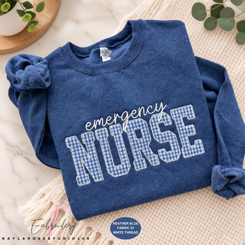 May include: A heather blue long-sleeve sweatshirt featuring the word emergency in white script and the word NURSE in a blue and white gingham pattern with white stitched outlines. The fabric has a textured, washed-out appearance.