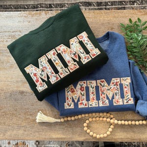 May include: Two sweatshirts with the word "MIMI" embroidered in floral fabric. The sweatshirts are in dark green and blue.