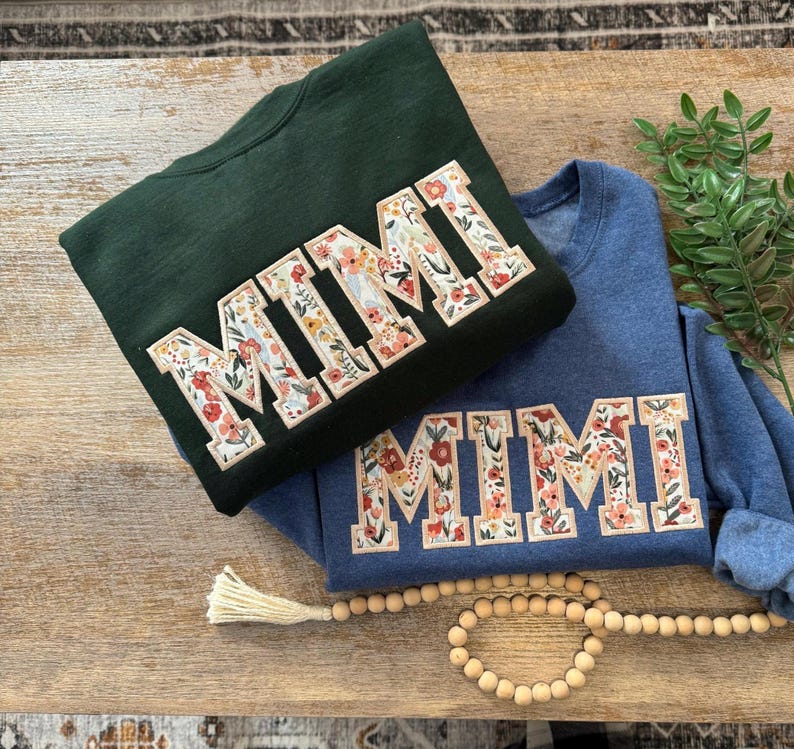 May include: Two sweatshirts with the word "MIMI" embroidered in floral fabric. The sweatshirts are in dark green and blue.