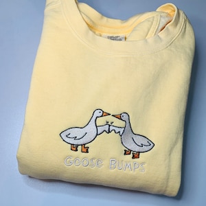 K&ouml;nnte beinhalten: Ein gelber Pullover mit einem wei&szlig;en gestickten Design aus zwei G&auml;nsen, die sich gegen&uuml;berstehen, mit dem Text "Goose Bumps" darunter.