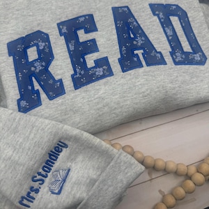 May include: A grey sweatshirt featuring the word "READ" in large, blue letters with a floral pattern. The sleeve cuff has the text "Mrs. Standley" and a small book illustration, both in blue. A wooden bead garland is visible.