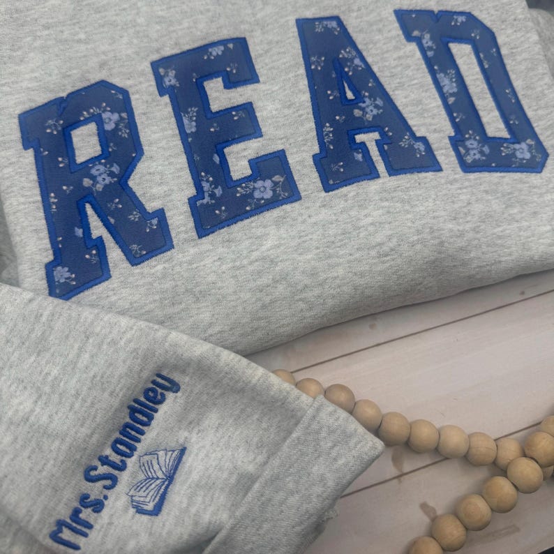 May include: A grey sweatshirt featuring the word "READ" in large, blue letters with a floral pattern. The sleeve cuff has the text "Mrs. Standley" and a small book illustration, both in blue. A wooden bead garland is visible.