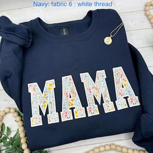 May include: Navy blue sweatshirt with the word "MAMA" embroidered in large, white-outlined letters filled with a floral pattern. The sweatshirt has a crew neck and long sleeves. A gold pendant necklace is visible.
