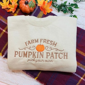 May include: A beige sweatshirt with the text "Farm Fresh Pumpkin Patch pick your own" embroidered in brown. The text is surrounded by a brown swirl design and a small orange pumpkin.