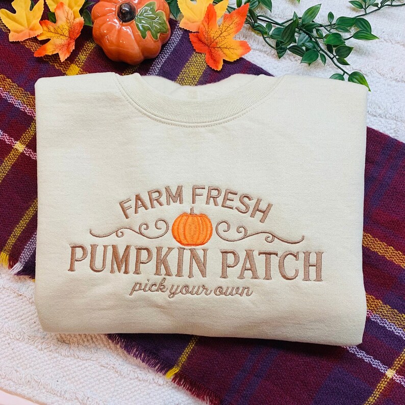 May include: A beige sweatshirt with the text "Farm Fresh Pumpkin Patch pick your own" embroidered in brown. The text is surrounded by a brown swirl design and a small orange pumpkin.