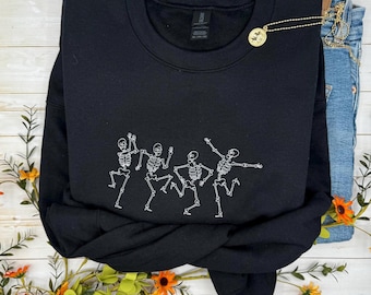 May include: A black sweatshirt featuring a white embroidered design of dancing skeletons. The crew neck sweatshirt is folded on top of a pair of blue jeans. A gold necklace with a pendant is on the sweatshirt.