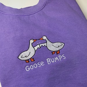 K&ouml;nnte beinhalten: Lilafarbener Pullover mit einem wei&szlig;en gestickten Design aus zwei sich gegen&uuml;berstehenden G&auml;nsen mit dem Text "Goose Bumps" darunter.
