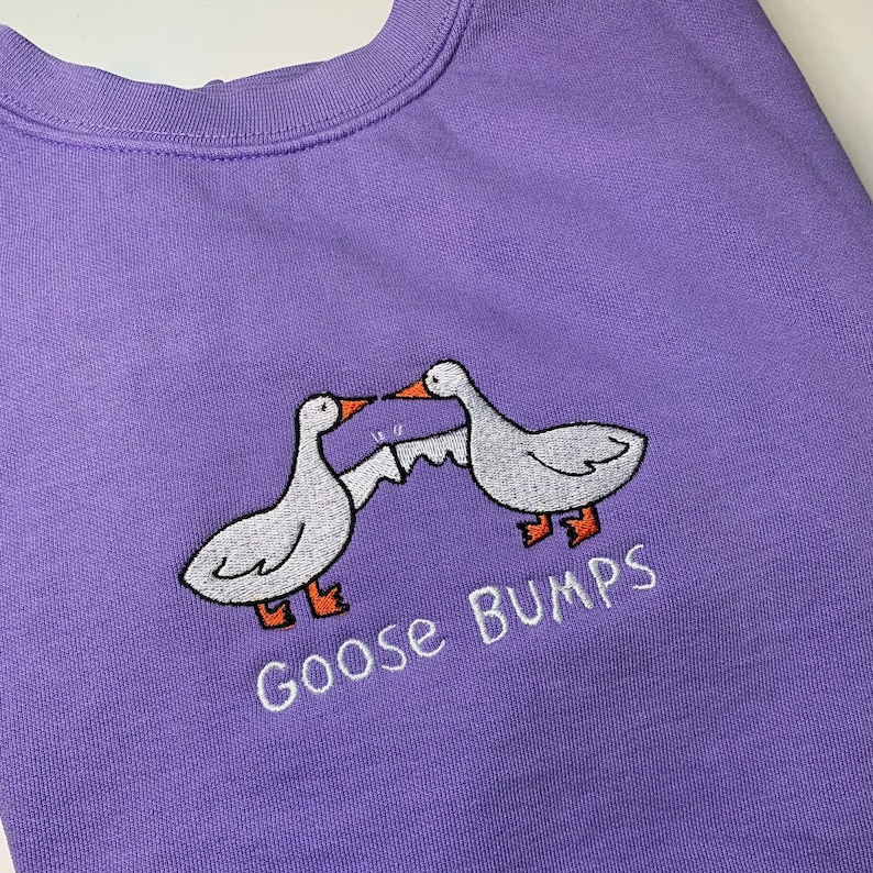 K&ouml;nnte beinhalten: Lilafarbener Pullover mit einem wei&szlig;en gestickten Design aus zwei sich gegen&uuml;berstehenden G&auml;nsen mit dem Text "Goose Bumps" darunter.