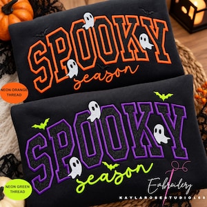 May include: Two black sweatshirts featuring embroidered text. The top sweatshirt has orange block letters reading SPOOKY and cursive text reading season, with white ghost and black bat accents. The bottom has purple and black text.
