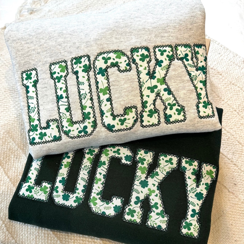 May include: Two sweatshirts with the word "LUCKY" written in large, bold letters. The letters are outlined in black and filled with a green shamrock pattern. The top sweatshirt is gray and the bottom sweatshirt is black.