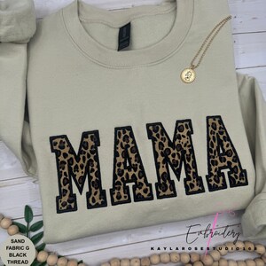 May include: A sand-colored sweatshirt with the word "MAMA" embroidered in large, block letters with a leopard print design and black outline. A gold necklace with a pendant rests on the sweatshirt. The text "SAND FABRIC G BLACK THREAD" is visible.