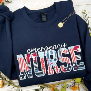 Op de afbeelding: Een marineblauw sweatshirt met ronde hals en de witte tekst "emergency" boven het woord "NURSE" in grote blokletters. De letters zijn gevuld met een patriottisch rood, wit en blauw patroon van sterren en strepen.